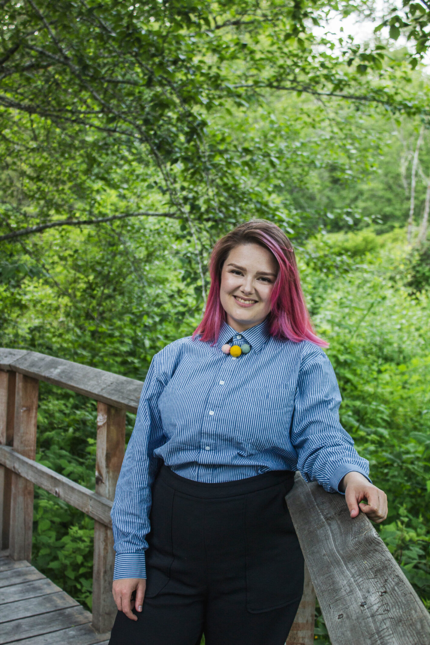 A femme person in a lush forest smiles warmly. They have pink hair and are dressed semi-formally in a blue buttonup dress-shirt, and black high-waisted dress pants.